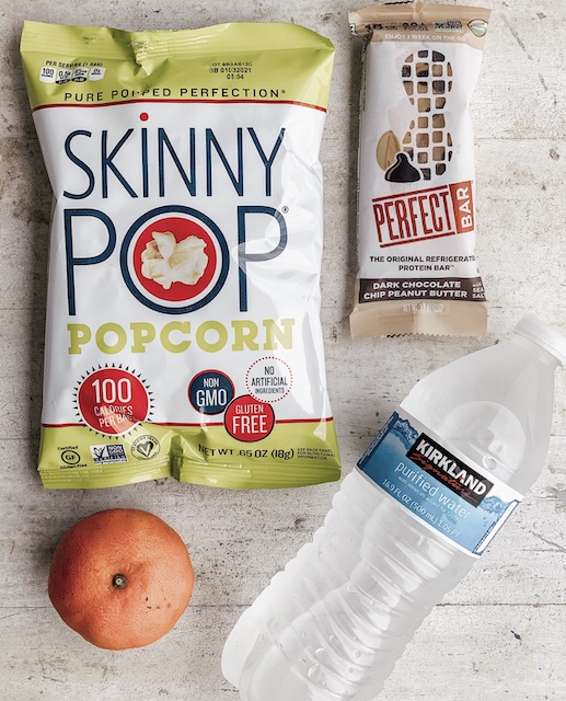 A snack consisting of popcorn, an orange, a bar and a bottle of water sitting on a table.