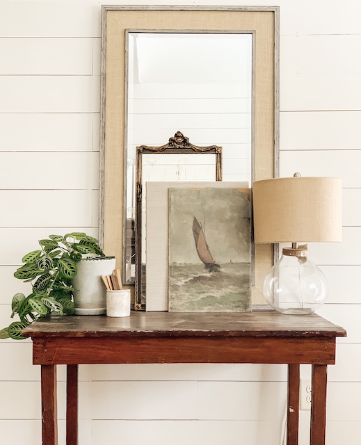 An antique table with a lamp, mirror, oil painting, and plant.