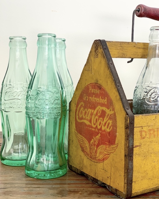 Three old coke bottles sitting next to a yellow coke caddy on a table.