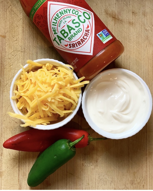 A lot of toppings for chili sitting on a cutting board including cheese, hot sauce, sour cream, and jalapeno peppers