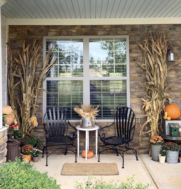 A wide view of a front porch with corn stalks and pumpkins and flowers