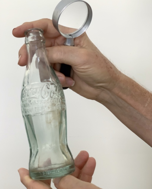 a man trying to date an old coca-cola bottle with a magnifying glass.