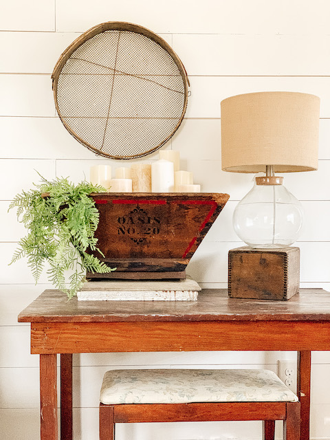 A table with a lamp and wooden creat and a grain sifter on the wall