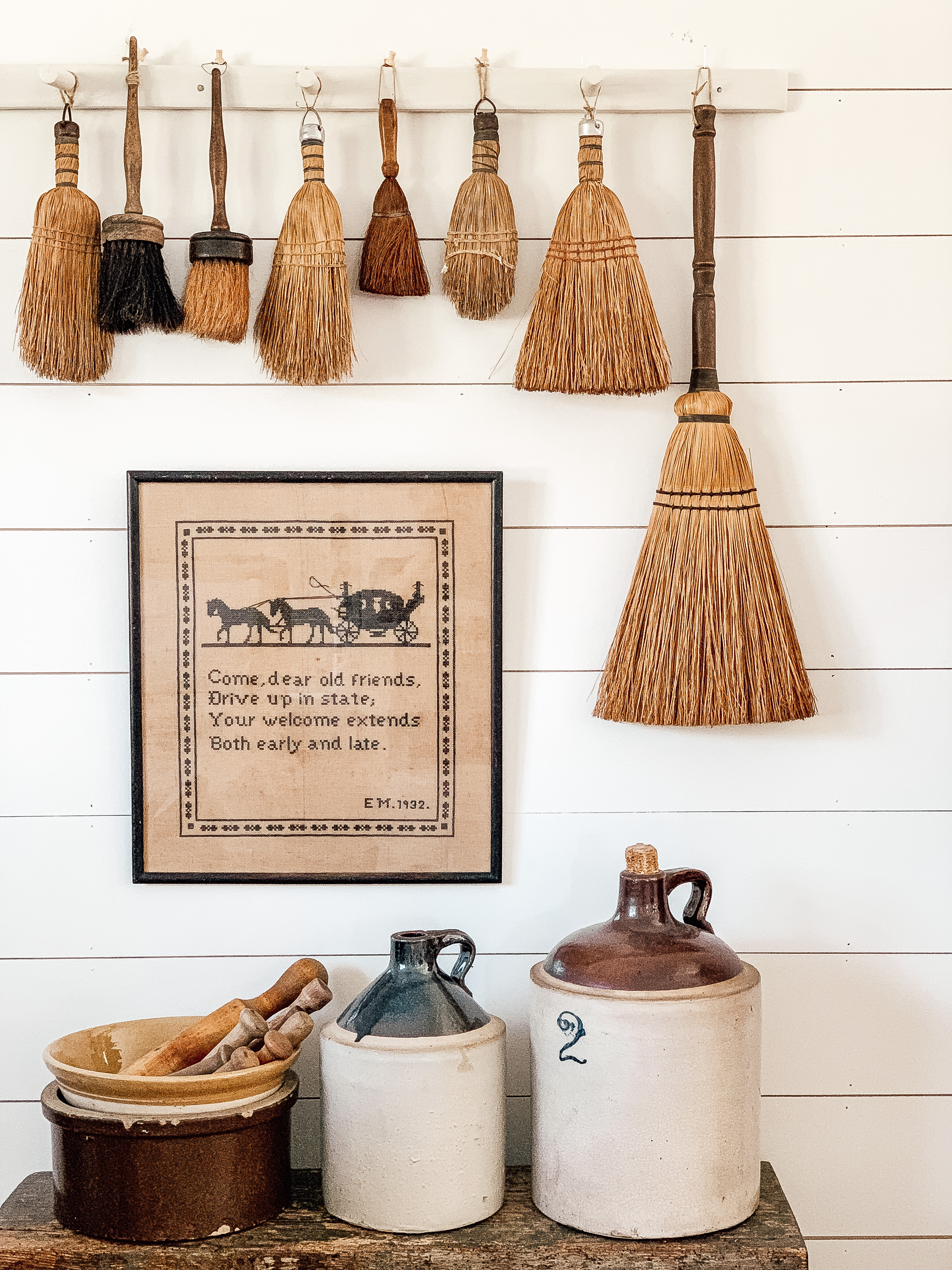 A vintage vignette with crocks, brooms, and a needle point